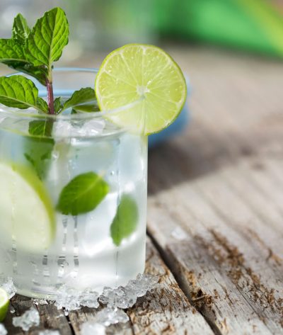 A mojito at the beach with lime and fresh mint with crushed ice on a rustic wood table with the ocean in the background.